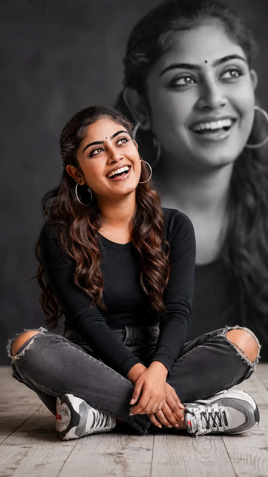 Smiling young woman with long wavy hair, sitting cross-legged on a wooden floor wearing a black long sleeve top and ripped jeans, with a large black and white blurred portrait of her laughing in the background.